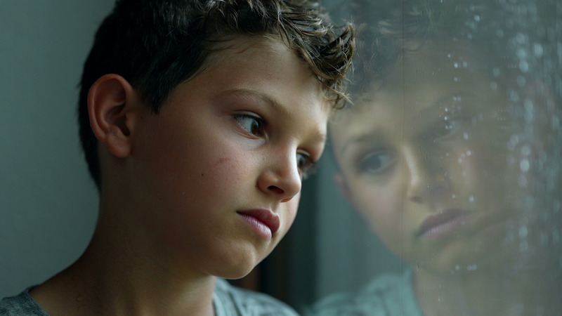 A young boy with short hair looks to the side and his reflection is visible in the window.